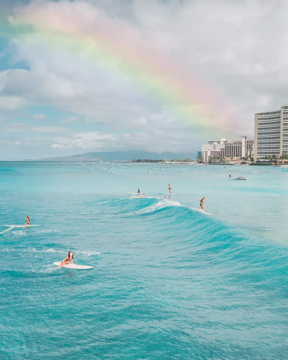 A rainbow arcing over the water off Oʻahu, surfers riding a long wave below it