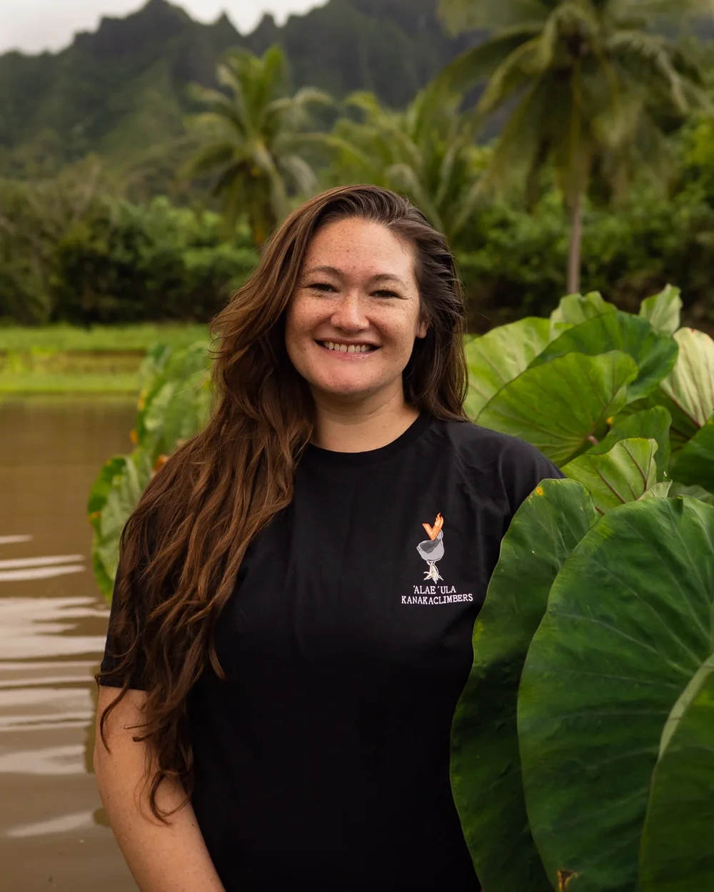 Kolealani “Kolea” Skye Razon-Olds standing in a loʻi, the ridge and palms behind her