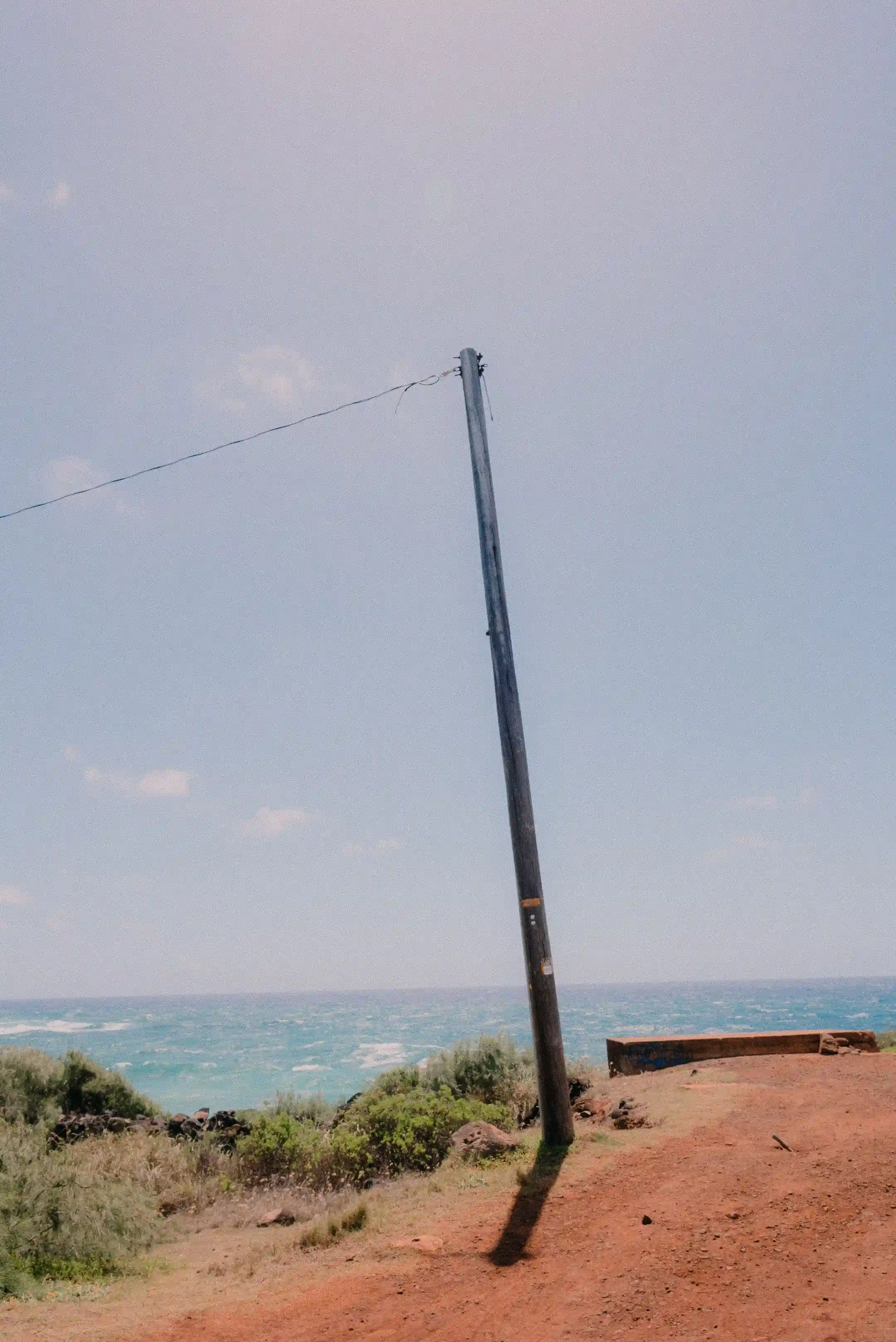 A lone utility pole leaning on a red dirt bluff above the ocean on a Hawaiʻi coastline