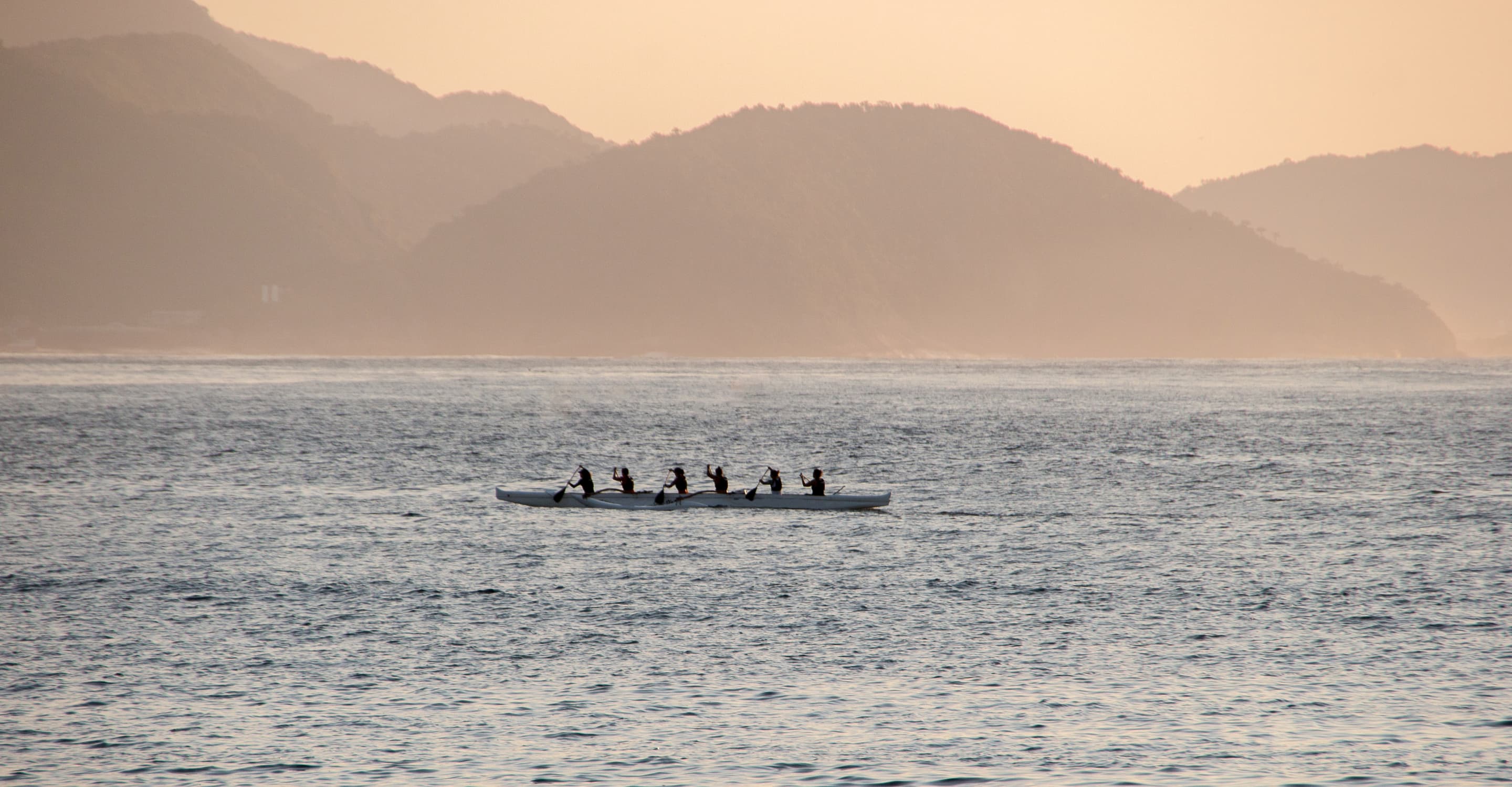 A six-person outrigger canoe crossing calm water below hazy ridgelines at sunset